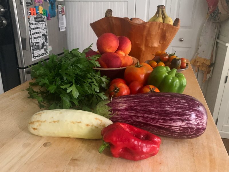 Atop the wooden tabletop of a kitchen island and within a variety of bowls sit parsley, tomatoes, eggplants, cucumbers, and peppers from the Bread and Roses CSA… plus some grocery store bananas and peaches.