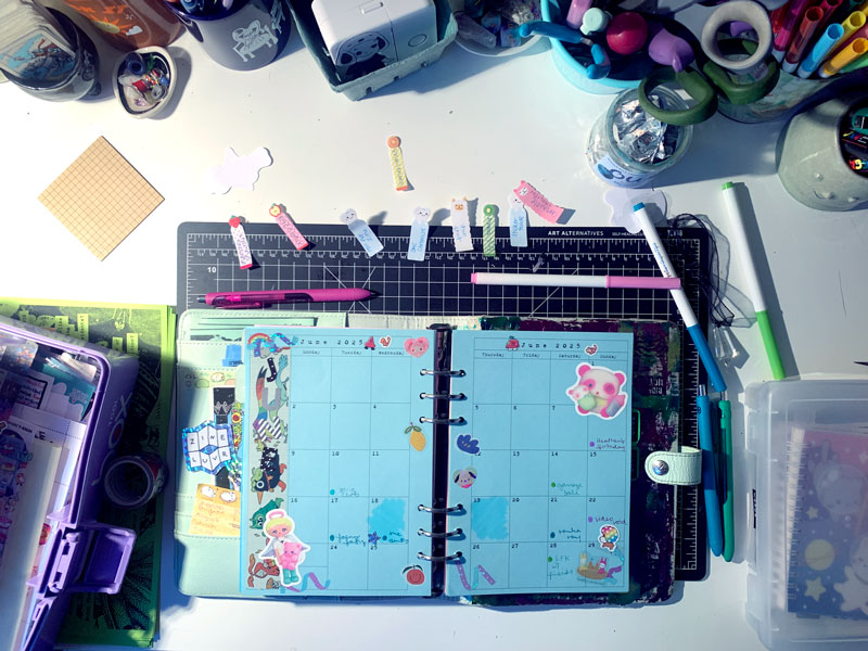 An overhead shot of a desk cluttered with cups of pens, scissors, markers, bits of foil, and mini tarot cards. In the center of the desk, ontop of a black gridded cutting mat is a mint green binder planner laying open and flat to a June calendar page printed on blue paper. The page is covered in stickers and scribbled notes of events, too small to make out in the image what they are.