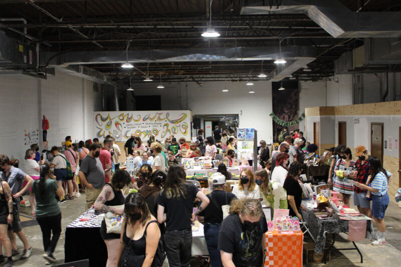 A zine fest: in a skatepark, the room is filled with tables from wall to wall where people wearing masks look at zines.
