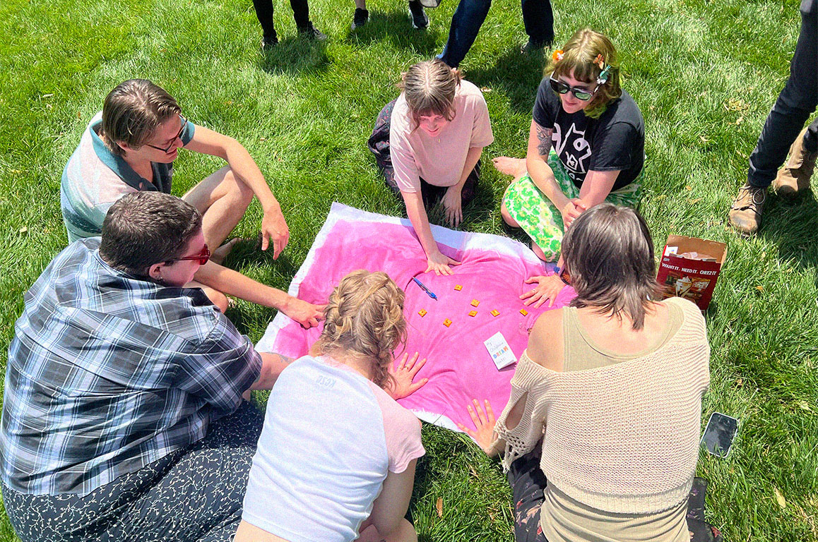 A bunch of zinesters gathered around a cloth treated with cyanotype. Their hands are on the cloth, as well as cheeze-its, a pen, and zines, to cast shadows on the fabric.