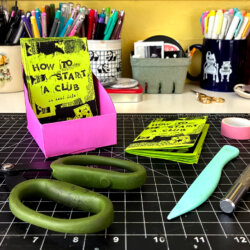 A pink paper zine holder and the supplies used to make it.
