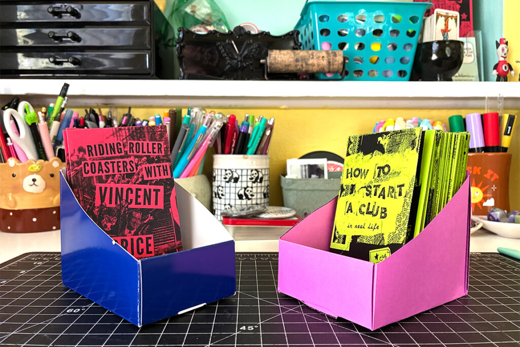 The finished zine holder next to the watch packaging that inspired the project, both containing zines.